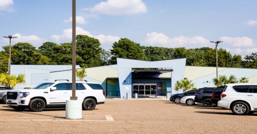 A light blue building with a modern design, surrounded by parked cars and trees under a partly cloudy sky.