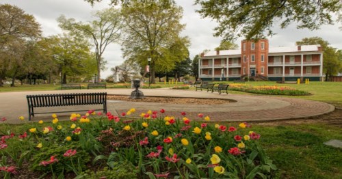 A park with colorful flower beds, benches, and a historic building in the background under a cloudy sky.