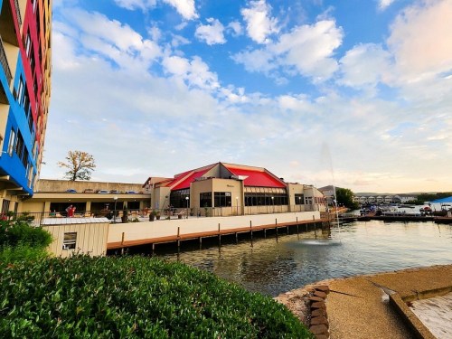 A waterfront building with a red roof, surrounded by water and greenery, under a blue sky with scattered clouds.