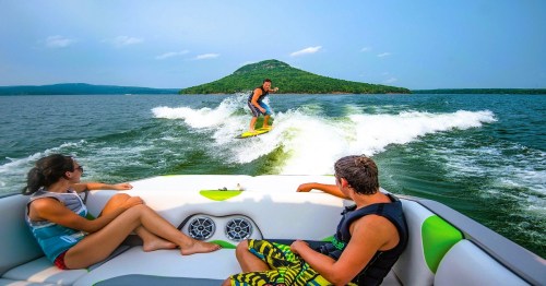 A person wakesurfing behind a boat, while two others relax on the boat, with a scenic island in the background.