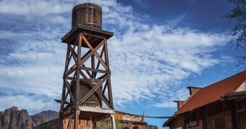 A wooden water tower stands tall against a blue sky with clouds, near rustic buildings and mountains in the background.