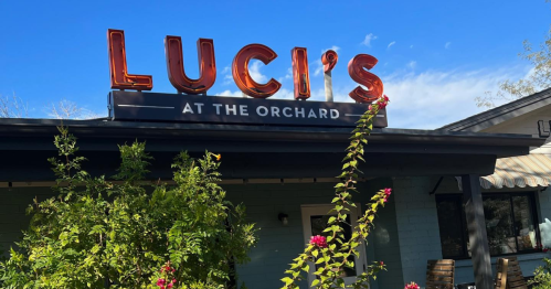 Sign for "Luci's at the Orchard" with greenery and blue sky in the background.