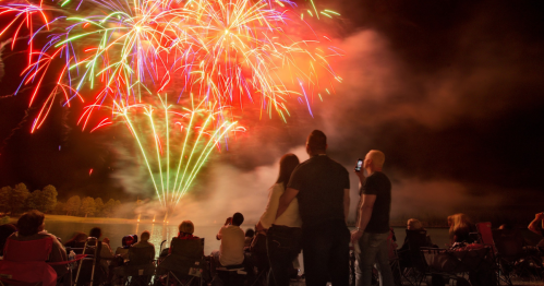 A crowd watches colorful fireworks explode over a lake at night, with people sitting in chairs and enjoying the display.
