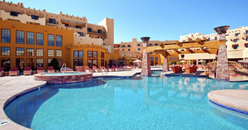 A sunny pool area with lounge chairs and a southwestern-style building in the background. Clear blue water reflects the sky.