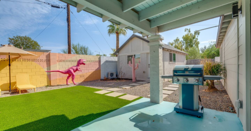 A backyard with a grill, artificial grass, and a pink dinosaur mural on the wall, surrounded by palm trees.