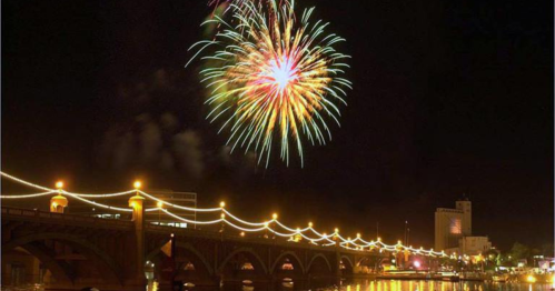 Colorful fireworks illuminate the night sky above a river, with a bridge and city lights reflecting in the water below.