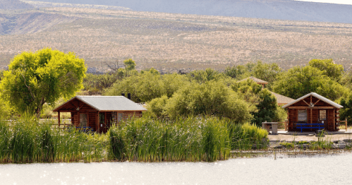 Two rustic cabins by a serene lake, surrounded by lush greenery and distant hills under a clear sky.