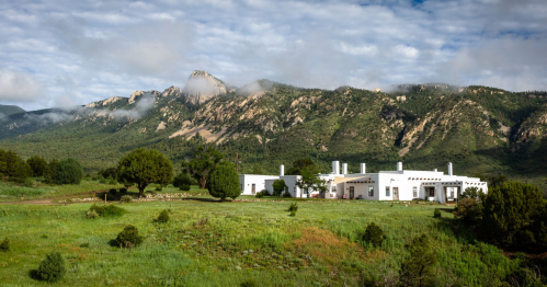 A white building surrounded by green fields, with mountains and clouds in the background.