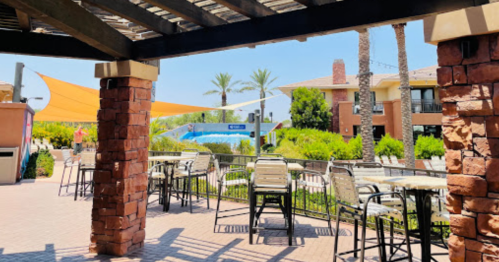 A sunny outdoor patio with tables and chairs, overlooking a pool and palm trees in a resort-like setting.