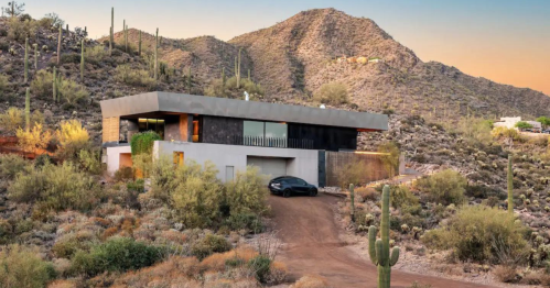 Modern house nestled in a desert landscape, surrounded by cacti and mountains, with a driveway leading to the entrance.