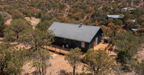 Aerial view of a rustic black cabin surrounded by trees and dry landscape, featuring a wooden deck.