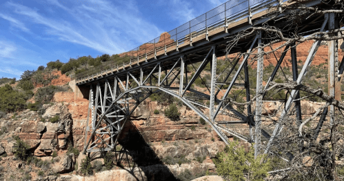 A metal arch bridge spans a rocky canyon, surrounded by greenery and red rock formations under a blue sky.