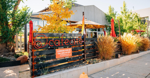 A cozy café with outdoor seating, surrounded by trees and colorful fall foliage, featuring a sign for curbside pickup.