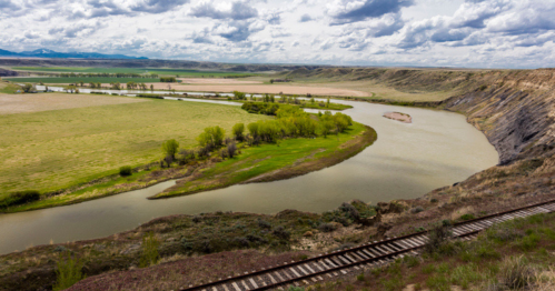 A winding river flows through green fields and hills under a cloudy sky, with a railway track in the foreground.