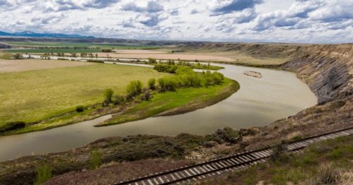 A winding river flows through green fields and hills under a cloudy sky, with a railway track in the foreground.