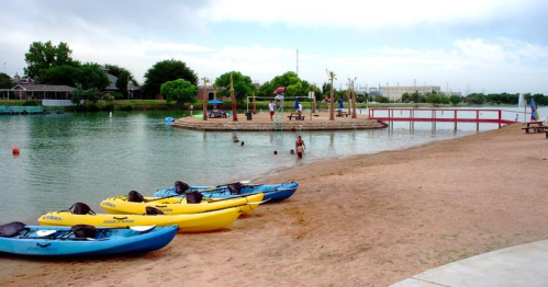 A sandy beach with yellow and blue kayaks, people swimming, and a playground on a small island in the background.
