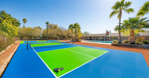 Bright outdoor tennis courts with green and blue surfaces, surrounded by palm trees and a pool area in the background.