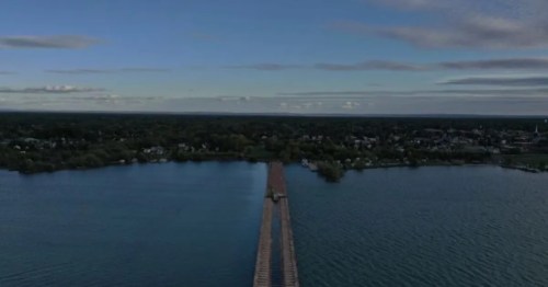 Aerial view of a long bridge extending over a calm body of water, surrounded by greenery and a clear sky.