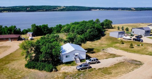 Aerial view of a white house near a lake, surrounded by trees and open land, with several parked cars nearby.