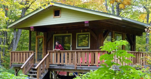 A cozy wooden cabin surrounded by trees, with a man in a flannel shirt standing on the porch.