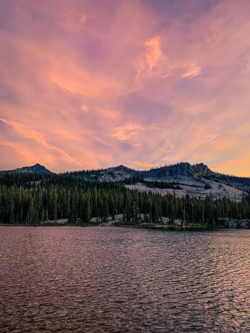A serene lake reflects a colorful sunset, surrounded by mountains and dense evergreen trees.