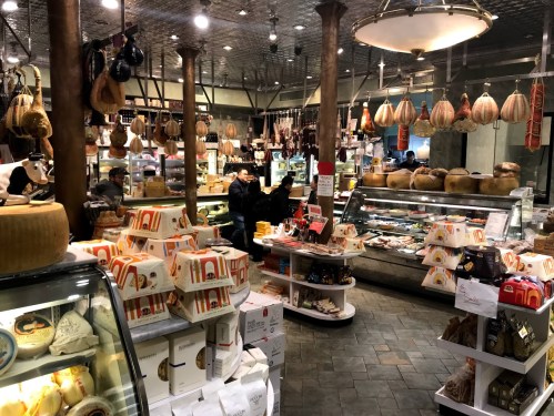A bustling market interior filled with various cheeses, meats, and packaged goods, with customers browsing and shopping.