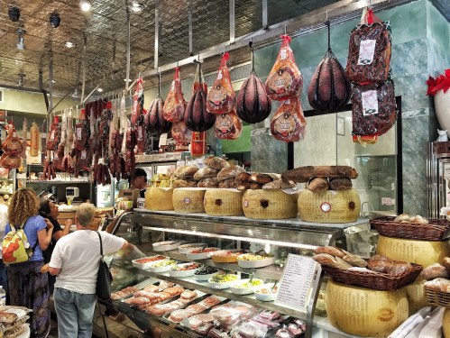 A bustling deli with hanging cured meats, various breads, and prepared dishes displayed in glass cases.