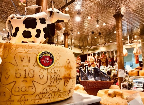 A cheese wheel with a cow figurine on top, surrounded by various cheeses and cured meats in a market setting.