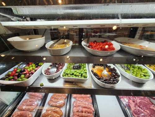 A display case filled with various salads, olives, and meats, showcasing colorful dishes in white bowls.