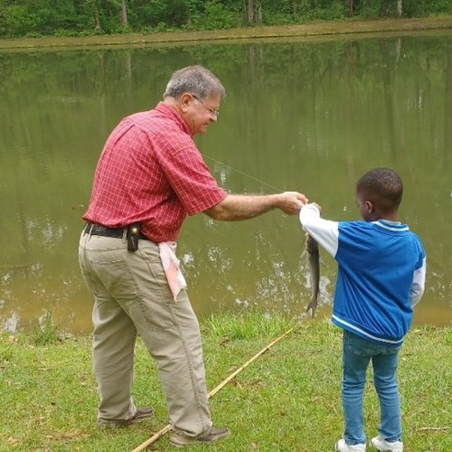 An adult man and a child stand by a lake, with the man holding a fish caught on a fishing line.