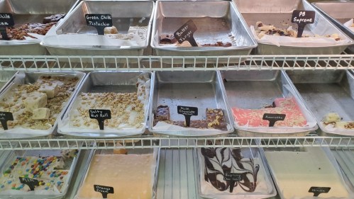 A display of various fudge flavors in metal trays, labeled with black signs, including peanut butter and raspberry cream.