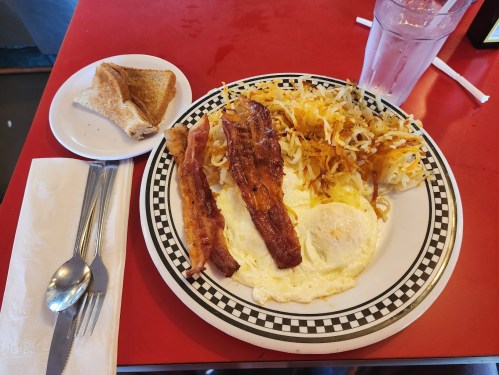 A plate of breakfast featuring eggs, bacon, hash browns, and a slice of toast, with a glass of water on the side.