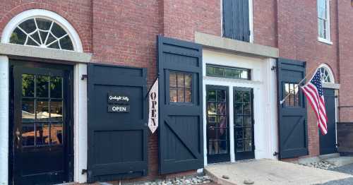 A historic brick building with open black doors, an "OPEN" sign, and an American flag displayed outside.