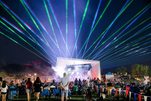 A lively outdoor concert at night with colorful laser lights and a crowd enjoying the performance.