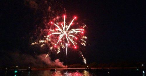 Colorful fireworks burst over a dark lake, reflecting vibrant lights on the water's surface.