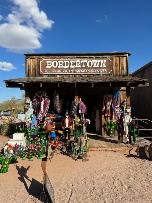 A rustic shop named "Bordertown" selling dolls, Mexican imports, and knives, adorned with colorful decorations outside.
