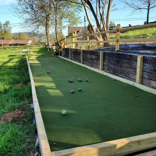 A grassy bocce ball court with several balls scattered on the surface, surrounded by trees and a wooden fence.