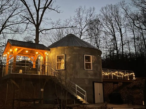 A unique round house with a deck, illuminated by string lights, surrounded by trees at dusk.