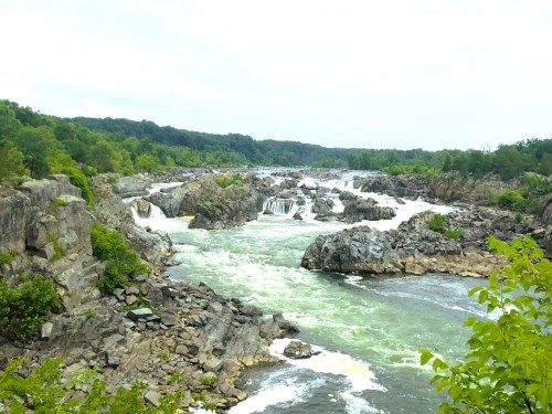 A scenic view of a river with rocky banks and cascading waterfalls, surrounded by lush greenery under a cloudy sky.