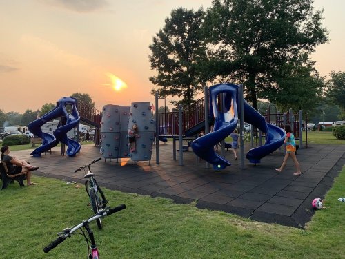Children play on a colorful playground with slides and climbing walls, set against a sunset sky. Bikes are nearby.