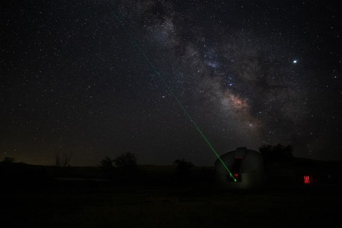 A green laser beam points towards the Milky Way, illuminating a dark sky above a small observatory.
