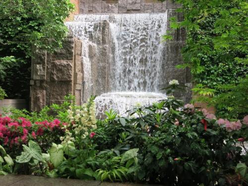 A serene waterfall cascades over rocks, surrounded by vibrant green foliage and colorful flowers.