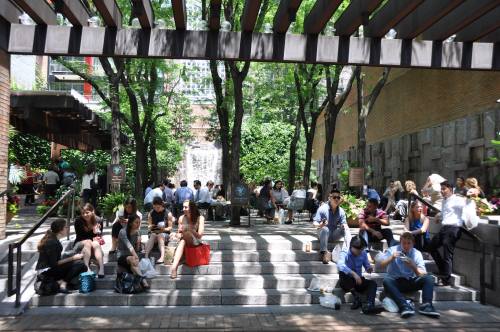 A lively urban park with people sitting on steps, surrounded by trees and greenery, enjoying the sunny day.
