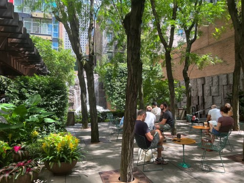 A serene outdoor space with trees, plants, and people sitting at tables, enjoying a sunny day in a garden setting.