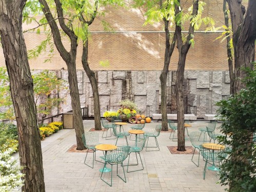 A serene outdoor space with green chairs, tables, and autumn decorations among trees and textured stone walls.