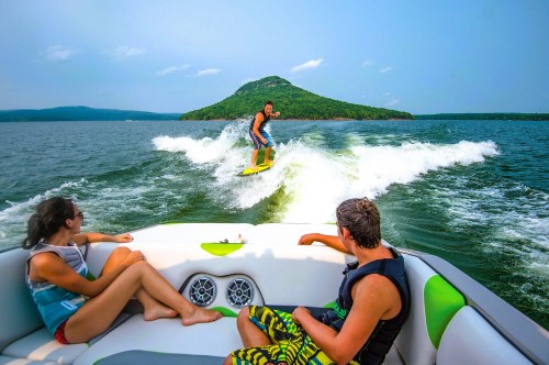 A person surfing behind a boat on a lake, with two people relaxing on the boat and a scenic mountain in the background.