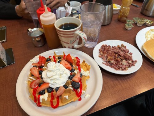 A plate of waffles topped with strawberries, blueberries, whipped cream, and syrup, alongside a side of hash and coffee.