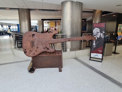 A large, rusty guitar sculpture stands in an airport, next to a Rock & Roll exhibit banner.