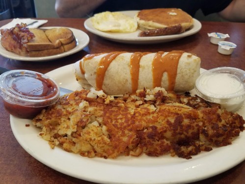 A plate with a burrito drizzled with sauce, crispy hash browns, and two dipping sauces, with pancakes and toast in the background.
