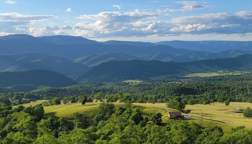 A scenic view of rolling green hills and mountains under a blue sky with fluffy clouds. A small building is visible in the foreground.